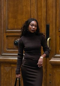 Bruine geribbelde coltrui en nauwsluitende rok, beide in diepbruin. Accessoires omvatten gouden oorbellen en een zwarte handtas. Houten achtergrond.