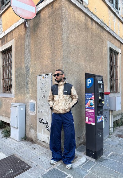 Chaqueta de camuflaje de North Face con acentos negros, pantalones anchos azul marino y zapatillas grises, de pie junto a un parquímetro en una esquina de la calle.