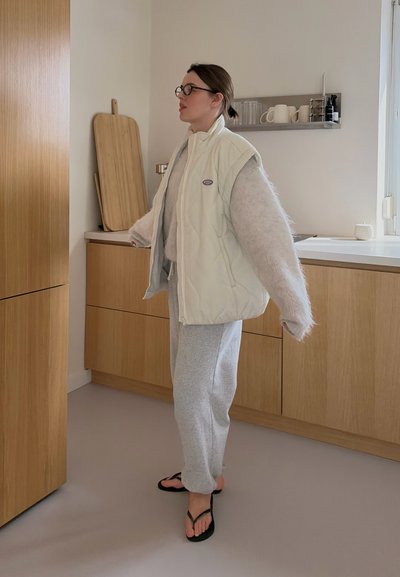 Mujer con gafas que lleva un chaleco blanco, un suéter peludo, pantalones deportivos grises y chanclas negras, de pie en una cocina moderna de madera.