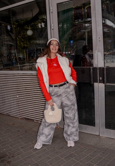 Llevando una camisa de manga larga roja, un chaleco de piel sintética blanca, pantalones grises con patrón de mármol y sosteniendo un bolso crema; zapatos de plataforma con acentos.