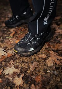 Fekete kültéri cipők, texturált felülettel, kiemelkedő talpmintázattal és rugalmas fűzőkkel, avarral borított talajon elhelyezve.