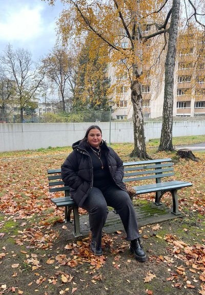 Una persona con una chaqueta acolchada negra está sentada en un banco de parque verde rodeado de hojas caídas de color naranja y marrón, con árboles al fondo.
