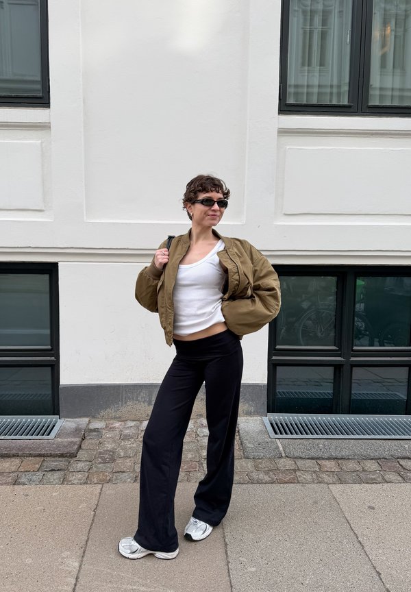 Mujer con chaqueta marrón, camiseta blanca, pantalones negros y gafas de sol, de pie en la acera frente a un edificio blanco con grandes ventanas.