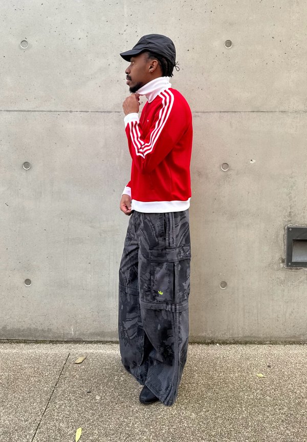 Man die een witte kraag onder een rood jasje met witte strepen aanpast, draagt een zwarte pet en losse donkere cargobroek, staand tegen een betonnen muur.