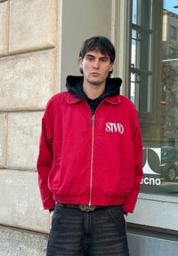 Κόκκινο φούτερ με φερμουάρ και ελαστικές μανσέτες, με λογότυπο "STWD" σε λευκό. Φορεμένο πάνω από μαύρη μπλούζα με κουκούλα, συνδυασμένο με σκούρα τζιν. Αστικό σκηνικό.