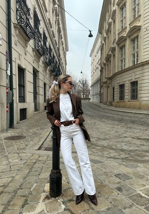 Blazer marrón oversize, camiseta blanca, jeans anchos blancos, cinturón marrón y tacones marrones de punta, de pie en una calle de adoquines.