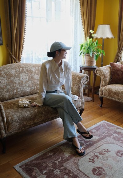 Mujer con ropa informal y gorra sentada en un sofá con estampado en una sala de estar luminosa de estilo vintage con planta en maceta y lámpara de pie cerca.