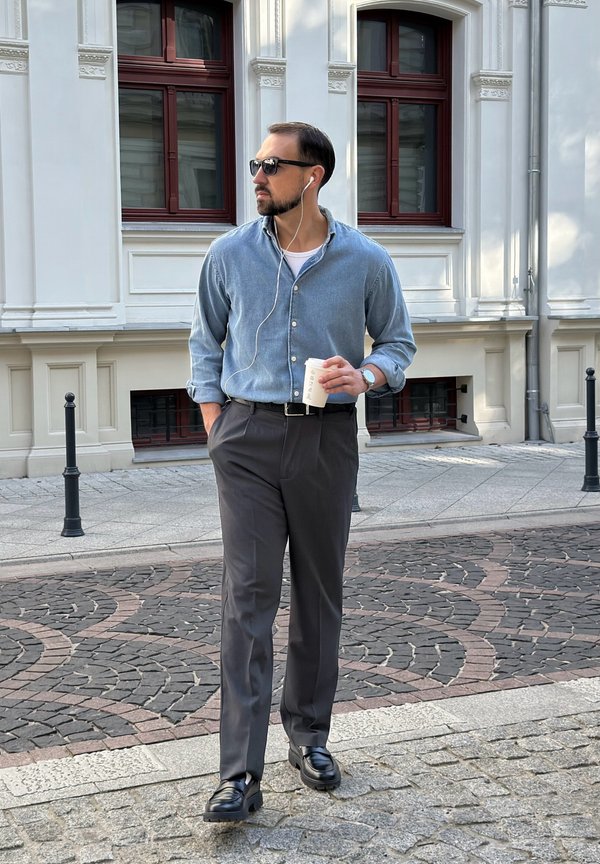 Hombre con gafas de sol y auriculares camina por una calle adoquinada con patrón sosteniendo una taza de café, cerca de un edificio blanco con grandes ventanas.