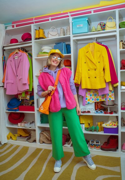 Mujer con ropa informal colorida con pantalones verdes, chaleco rosa, gorra amarilla y bolso naranja, de pie sonriendo frente a un armario con ropa y zapatos.