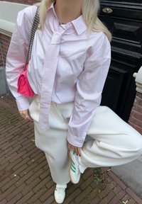 Prúžkovaná ružová košeľa na zapínanie s uzlovým detailom, biele široké nohavice a biele tenisky so zelenými akcentmi. Malá ružová kabelka s reťazovým remienkom.