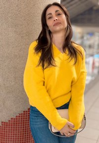 Femme aux longs cheveux bruns portant un pull jaune vif et un jean bleu, tenant un sac beige, appuyée contre un mur carrelé.