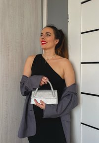 Γυναίκα με μαύρο φόρεμα ενός ώμου και γκρίζο σακάκι κρατάει ασημένια τσάντα, χαμογελώντας με κόκκινο κραγιόν, στέκεται μπροστά σε έναν τοίχο με ξύλινη και λευκή επένδυση.