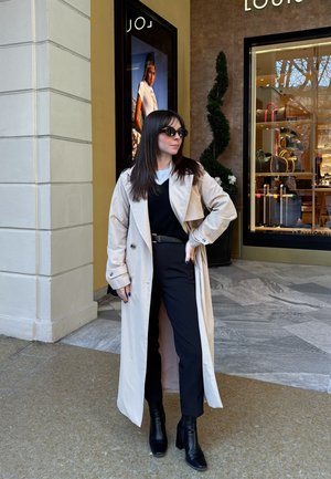 Trench coat beige, blusa negra, pantalones de tiro alto y botines negros de plataforma. Las gafas de sol y un cinturón complementan el conjunto, con un fondo de piedra.