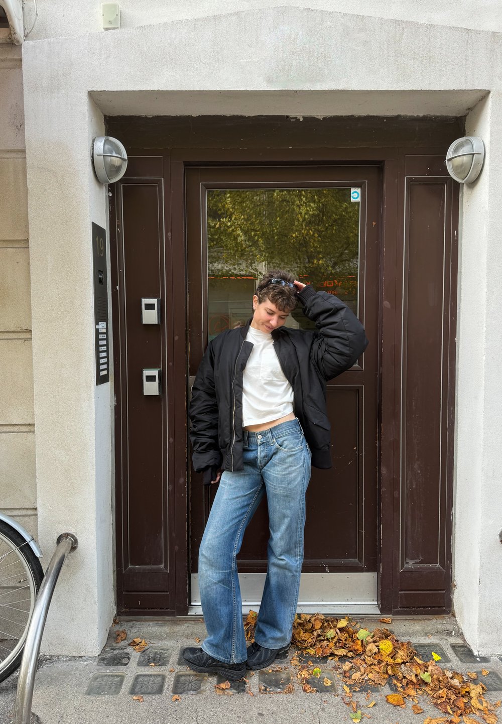 Joven con chaqueta negra, camiseta blanca y jeans de pie en la acera con hojas caídas frente a una puerta marrón con panel de entrada.