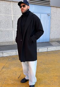 Μαύρο μάλλινο παλτό με σατέν γιακά, φορεμένο πάνω από ένα μαύρο ζιβάγκο. Ανοιχτό μπλε τζιν και μαύρα παπούτσια. Αξεσουάρ περιλαμβάνουν γυαλιά ηλίου και καπέλο.