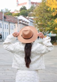 Vit texturerad jacka med fransade detaljer, kombinerad med en beige hatt med bred brätte som har ett brunt band, i en urban höstscen.
