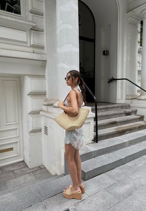 Bolso de paja trenzada con asas de cuero marrón, vestido ligero de estampado floral y cuñas beige sobre escalones de piedra cerca de un edificio blanco.