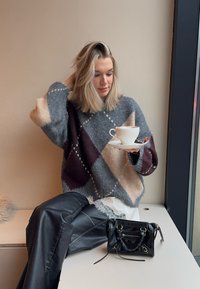 Ülenähtud halli, burgundia ja beeži värvi kudumissärk teemantmustritega, kombineeritud mustade nahkpükstega. Ühes käes hoitakse valget tassi taldrikul.