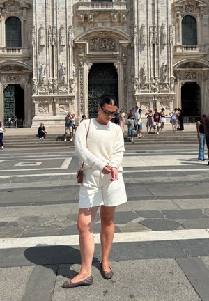 Suéter blanco de punto, pantalones cortos beige claros, zapatos planos marrones; de pie sobre un pavimento de piedra con un edificio histórico y personas al fondo.