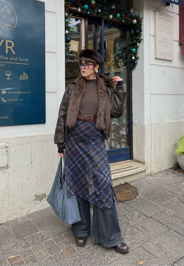 Vrouw met een bruine jas met bontkraag, een geruite rok over wijd uitlopende spijkerbroek, een baret, een bril en een grijze tas, staande voor een versierde cafédeur.