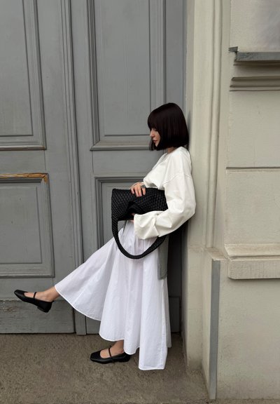 Mujer con falda y blusa blanca apoyada en la pared, sosteniendo un bolso negro tejido, con una pierna extendida, cerca de una puerta con paneles grises.