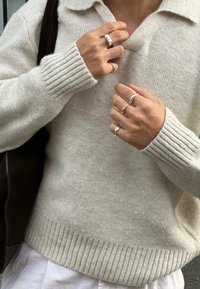 Stickad, beige tröja med ribbade muddar och nederkant, i v-ringning och med krage. Fyra silverringar på händerna, slät yta, mjuk uppenbarelse.