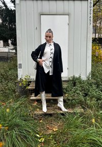 Svart lång kappa, vit skjorta med svarta rosetter, mörka byxor och vita klackstövlar. Står på trätrappor i ett grönskande område.