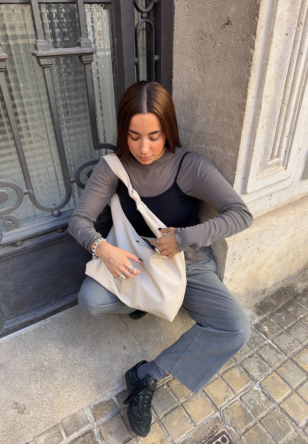 Beige tote bag with a textured pattern. The person is wearing a grey long-sleeve top and black sports shoes, seated against a stone wall.