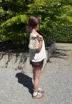 Mujer con top blanco y pantalones cortos negros de pie en un camino de grava, sosteniendo un bolso blanco con forma de concha sobre el hombro, con un fondo verde de hojas bajo la luz del sol.