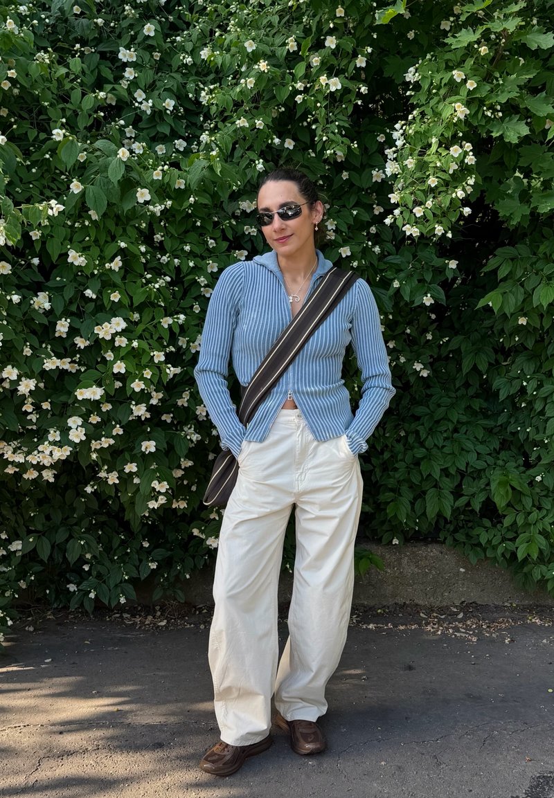 Femme portant des lunettes de soleil, une chemise rayée bleue, un pantalon blanc et un sac sombre, debout les mains dans les poches devant des buissons verts avec des fleurs blanches.