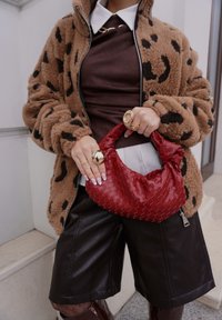 Veste en polaire marron clair et noire à motif, haut marron, short en cuir noir, sac à main rouge tissé avec un nœud, bagues en or, ongles longs.