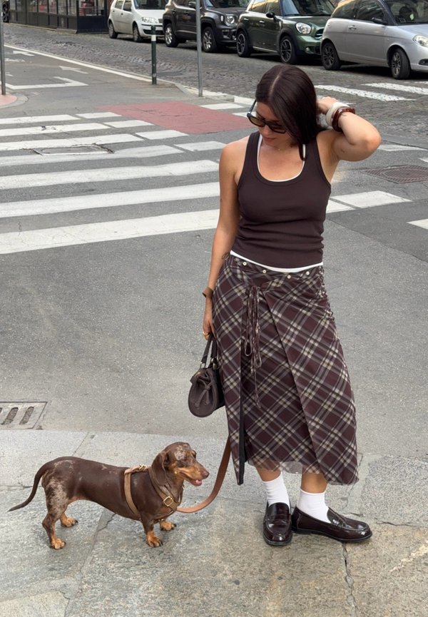 Mujer con gafas de sol, top sin mangas y falda de cuadros, de pie en la acera sosteniendo la correa de un dachshund marrón cerca de un paso de peatones con coches estacionados.