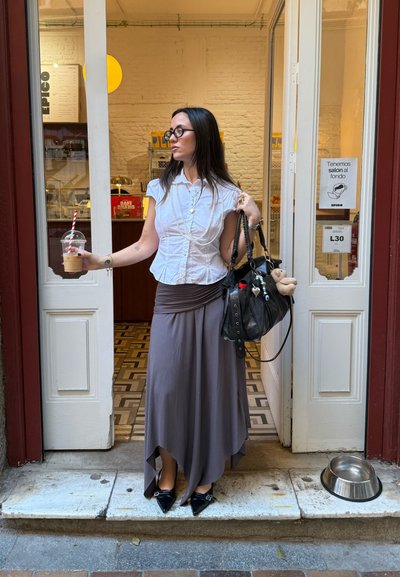 Mujer con una blusa blanca de manga corta y una falda maxi gris, sosteniendo una bebida, con un bolso negro decorado. Salen de un café.