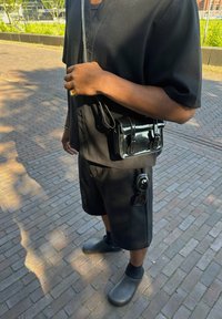 Camicetta nera a maniche corte, pantaloni corti neri e zoccoli grigi. Una piccola borsa a tracolla nera lucida e occhiali da sole sono visibili in vita.