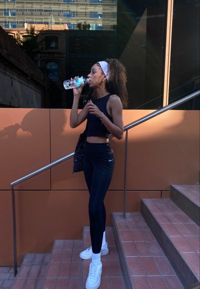 Top corto deportivo negro y leggings, zapatillas blancas y un bolso con estampado. Bebiendo de una botella de agua mientras está de pie en las escaleras.