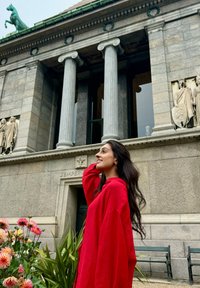 Een vrouw in een levendig rode, languit mouwen shirt staat nabij een stenen gebouw met zuilen en ingewikkelde beelden, omringd door kleurrijke bloemen.