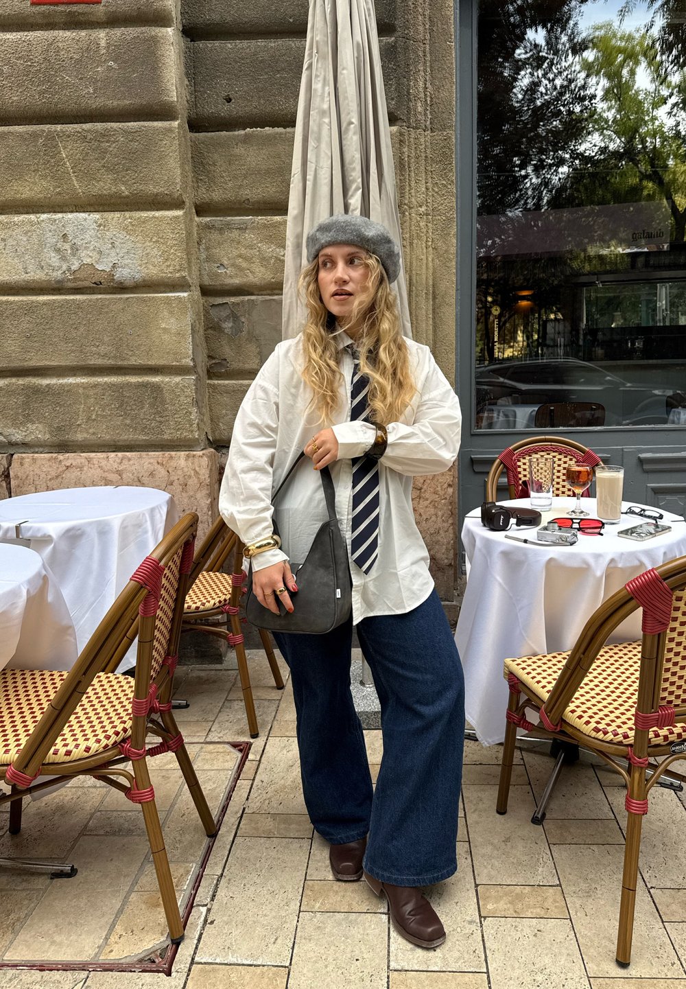 Modellen har på seg en hvit oversized skjorte, et stripete slips og blå brede jeans. Hun bærer en grå skulderveske og har på seg en grå hatt.