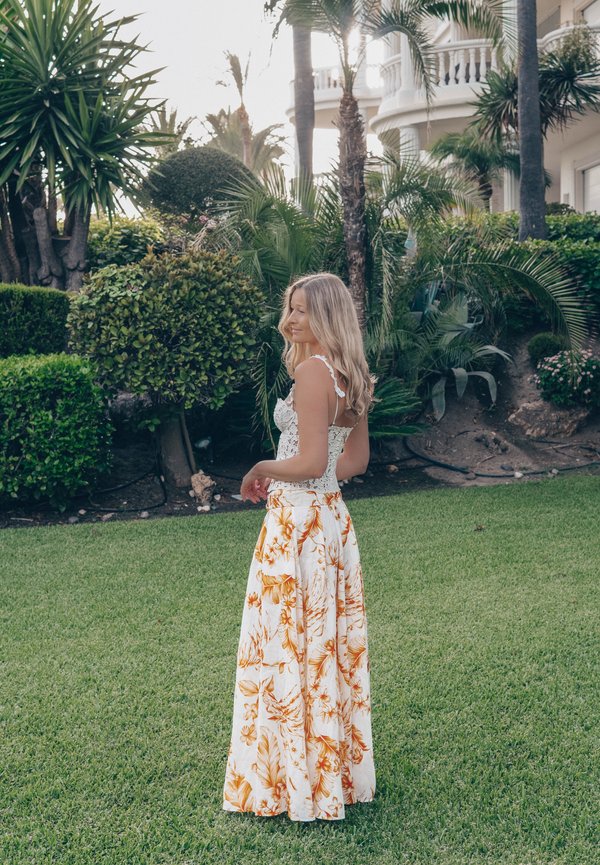 Kobieta w kwiecistej spódnicy i białym koronkowym topie stojąca na zielonym trawniku, w tle tropikalne rośliny i palmy.
