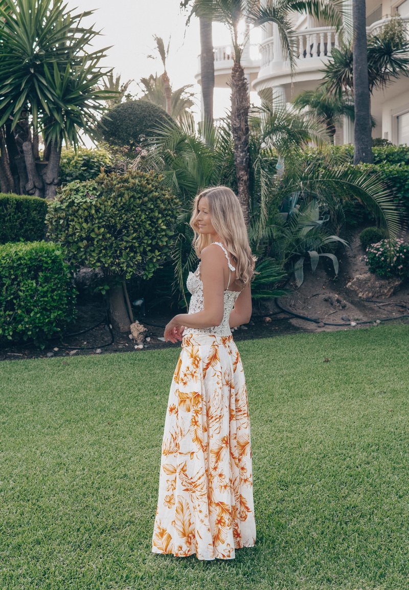 Mujer con falda de flores y top blanco de encaje, de pie sobre un césped verde con plantas tropicales y palmeras al fondo.