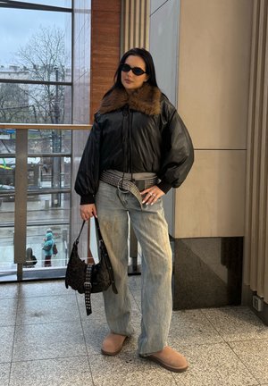 Mujer con chaqueta de cuero negra con cuello de piel, cinturón ancho, vaqueros sueltos, zapatos marrones, gafas de sol, sosteniendo un bolso negro en el interior junto a la ventana.