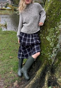 Chunky grå strikkegenser, rutete svart skjørt og grønne gummistøvler, står ved et tre nær vannet. Tekstur og mønstre synlige.