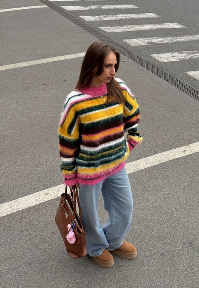 Suéter a rayas multicolor con tonos rosa, amarillo y verde, de textura suave. Combinado con jeans azul claro y botas marrones, sostiene un bolso marrón.