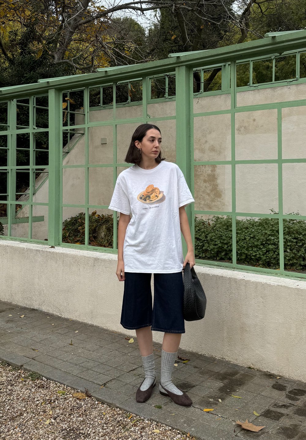 Hvid oversized t-shirt med et småkagegrafik, mørk denim culottes, grå stribede sokker og brune flade sko; holder en sort vævet taske.