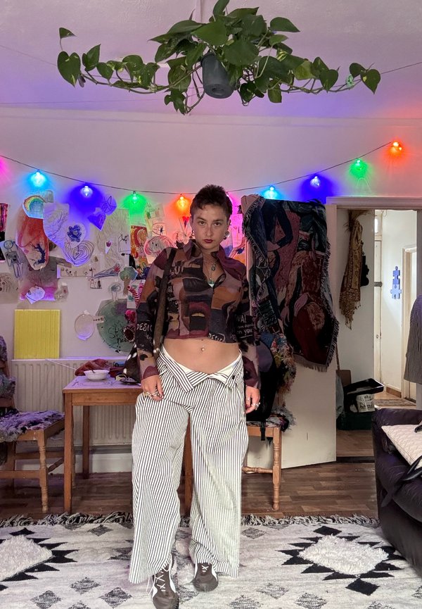 Person with short hair wearing a patterned cropped top and striped wide pants standing in a decorated room with colourful string lights and a hanging plant.