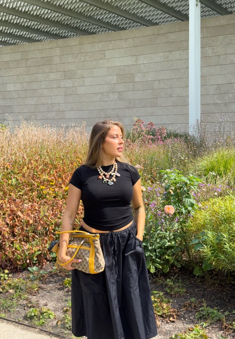 Woman in black top and skirt holding a yellow-accented bag stands in a garden with tall grass and flowers, looking to the side.