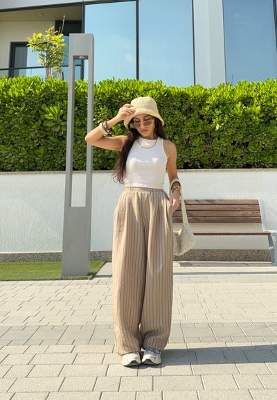 Pantalones anchos de rayas beige, camiseta sin mangas blanca, sombrero de paja estilo bucket, gafas de sol y bolso tejido. De pie en una zona exterior pavimentada.