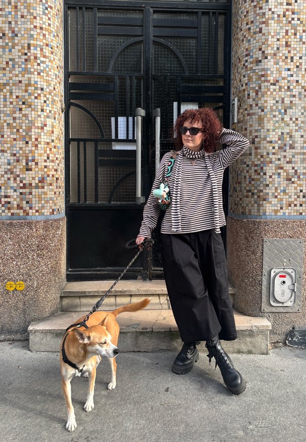 Moteris su garbanotais plaukais ir akiniais nuo saulės laiko šuns pavadėlį, stovėdama laiptinėse prie plytelių apdailos pastato su juodomis durimis.