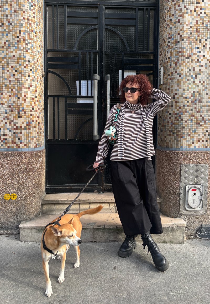 Kobieta z kręconymi włosami i okularami przeciwsłonecznymi trzymająca smycz psa, stojąca na schodach przed budynkiem z płytkami i czarnymi drzwiami.