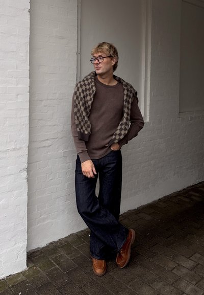 Joven con gafas, suéter marrón, bufanda a cuadros sobre los hombros, pantalones oscuros y zapatos marrones, apoyado contra una pared de ladrillos blancos.