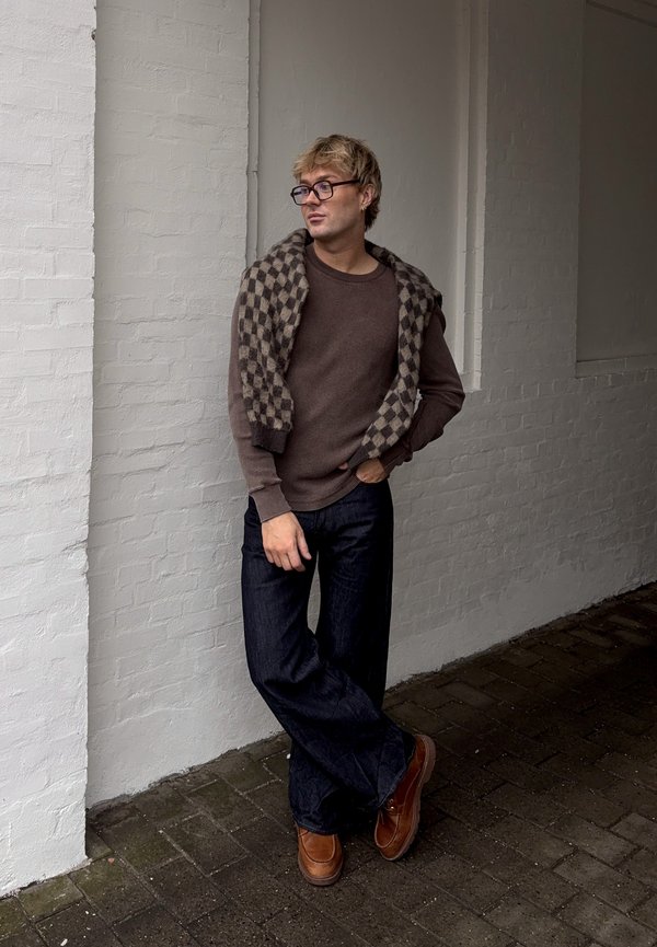 Ung mann med briller, brun genser, rutete skjerf over skuldrene, mørke bukser og brune sko, som lener seg mot en hvit murvegg.